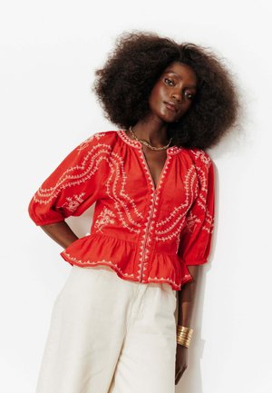 Femme aux cheveux bouclés volumineux portant une blouse rouge brodée avec des manches bouffantes et un pantalon blanc taille haute, debout sur fond blanc.