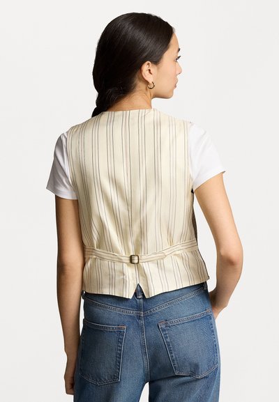 Gilet crème rayé avec un design ajusté, une sangle arrière réglable et une texture de tissu lisse, porté sur une chemise blanche et un jean bleu.