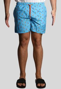 Shorts da bagno azzurri chiari con un motivo di uccelli colorati, vita elasticizzata e un laccetto arancione. Indossati con sandali neri.