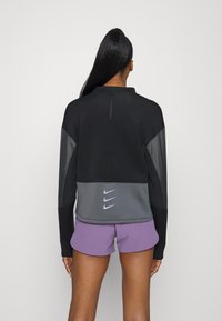 Maglietta nera a maniche lunghe con dettagli grigi, colletto con zip e inserti in rete. Presenta loghi Nike in azzurro chiaro sulla parte posteriore ed è abbinata a pantaloni corti viola.