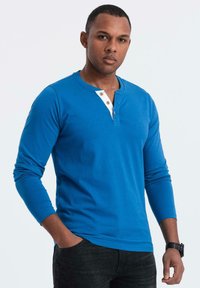 Langärmliges blaues T-Shirt mit weißem Kragen, das mit drei Holzknöpfen versehen ist. Getragen mit dunklen Jeans und einer schwarzen Armbanduhr.