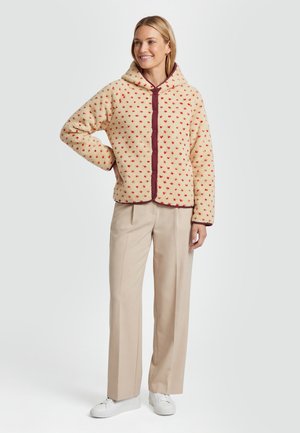 Smilende kvinde iført cremefarvet fleece-hoodie med røde detaljer, beige bukser med brede ben og hvide sneakers, står med den ene hånd på hoften.