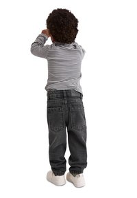 Tout petit avec des cheveux bouclés portant une chemise à rayures à manches longues, un jean noir et des baskets blanches, debout et dos tourné.