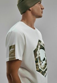 Biała bawełniana koszulka z zielono-brązowym wzorem czaszki w stylu kamuflażu z przodu, z teksturowaną metką z bambusa na rękawie.