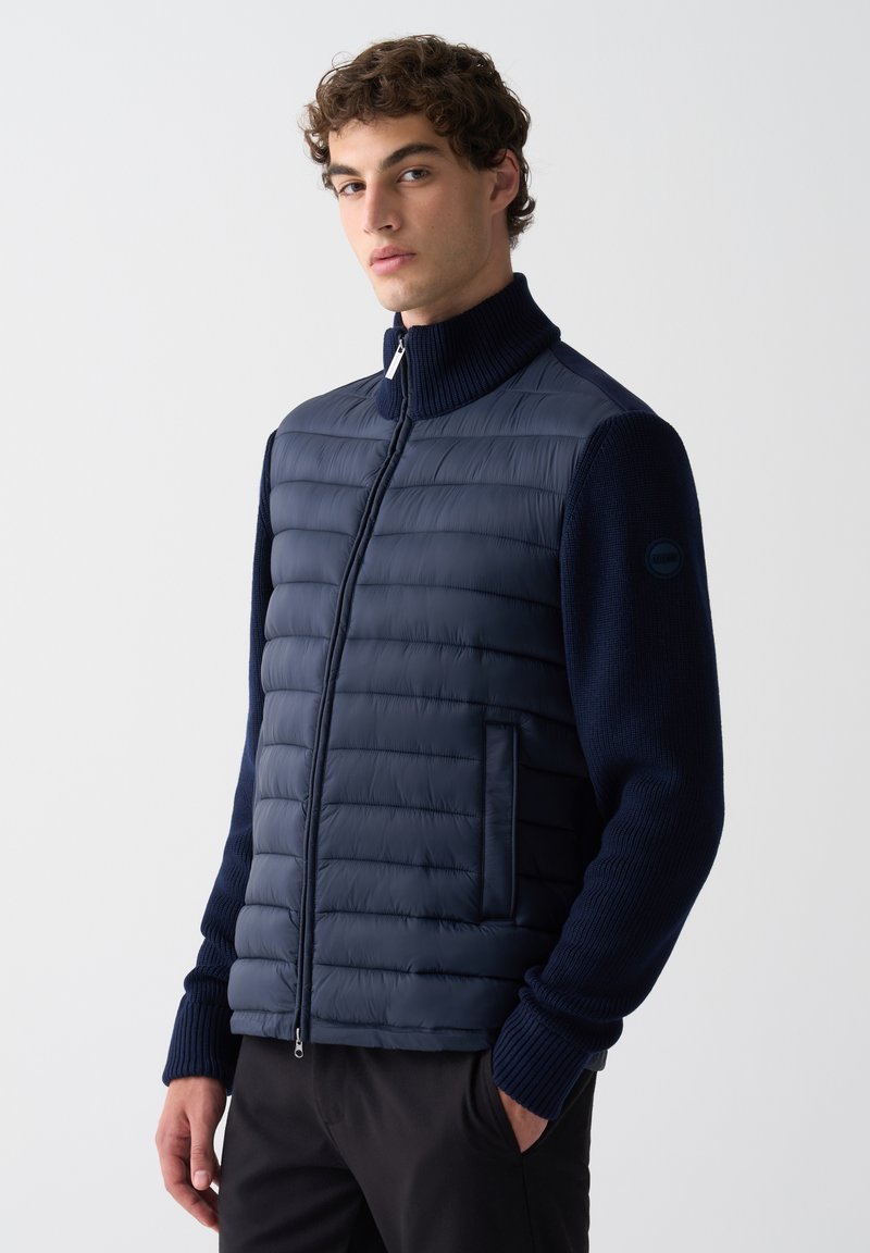 Mladý muž v námorní modrej prešívanej bunde s pletenými rukávmi, stojaci s ľavou rukou v vrecku pred bielym pozadím.