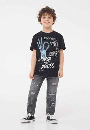 T-shirt nera con grafica di una mano scheletrica e testo bianco "CAMBIA LE REGOLE." Indossato con jeans grigi strappati e sneakers nere.