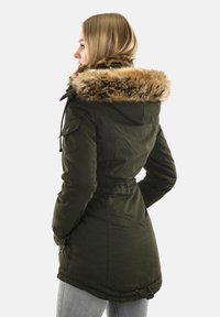 Eine Frau trägt einen olivgrünen Wintermantel mit einer mit Pelz gefütterten Kapuze, hat die Hände in den Taschen und blickt nach links vor einem schlichten Hintergrund.