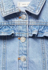 Ljusblå denimjacka med en frillad krage, silverfärgade tryckknappar, orange sömmar och en texturerad tyg.