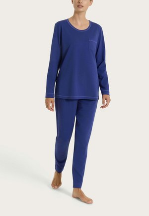 Persona che indossa una maglia a maniche lunghe blu scuro e pantaloni abbinati, camminando a piedi nudi su uno sfondo chiaro e neutro.