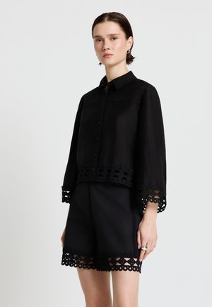 Frau trägt schwarzes Hemd mit Knöpfen und passende Shorts mit Spitzenbesatz an Ärmeln und Saum, vor einem schlichten hellen Hintergrund stehend.