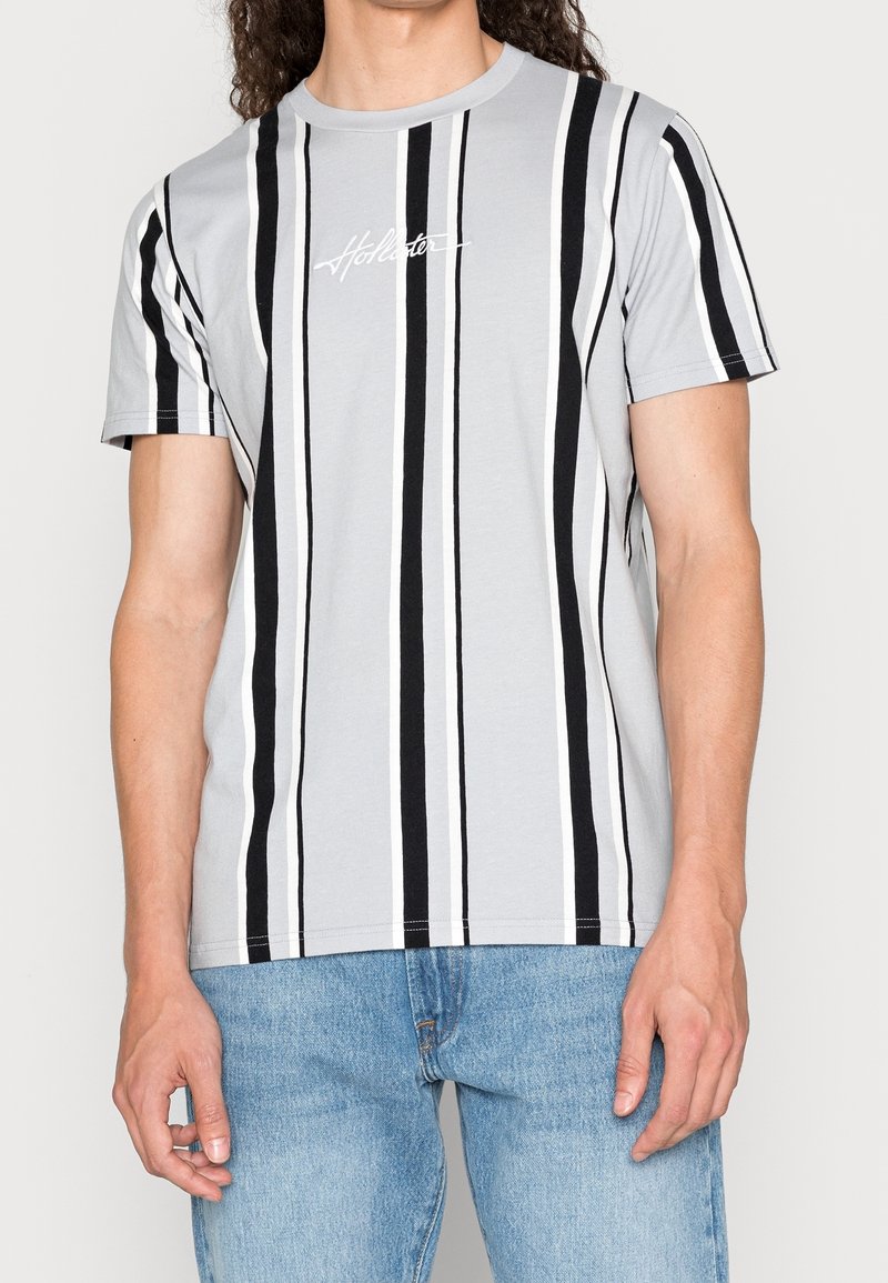 Grå kortärmad t-shirt med vertikala svart- och vitrandiga mönster och en vit broderad logotyp på bröstet.