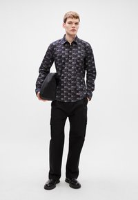 Πουκάμισο με κουμπιά σε σκούρες αποχρώσεις, με επαναλαμβανόμενο μοτίβο "HUG". Συνδυάζεται με μαύρο παντελόνι cargo και μαύρα παπούτσια, κρατώντας μια σκούρα τσάντα.