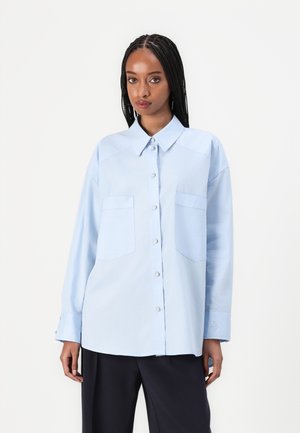 Lichtblauwe lange mouwen shirt met een puntige kraag, twee voorzakken, knoopsluiting en subtiele details bij de manchetten. Soepele stoftextuur.
