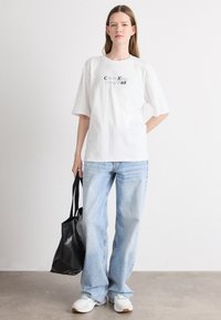 Witte katoenen t-shirt met korte mouwen en zwarte opdruk "Calvin Klein Since 1968," gecombineerd met lichtblauwe spijkerbroek en een zwarte tote bag.