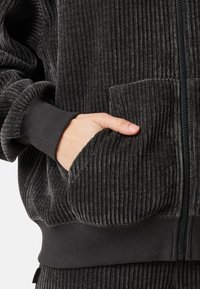 Schwarze gerippte Zip-Jacke mit Stehkragen, elastischen Bündchen und einer Vordertasche. Weicher, strukturierter Stoff mit einem gemütlichen Aussehen.