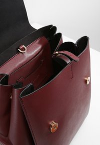 Sac à main en cuir rouge foncé avec une fermeture magnétique, ouvert pour montrer une poche intérieure et une sangle avec des ferrures dorées.