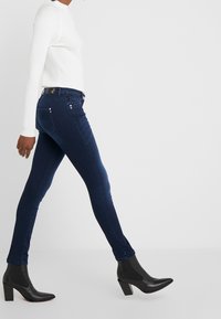 Jeans skinny azul oscuro con bolsillos traseros sutiles y accesorios plateados, combinados con una blusa blanca de manga larga con canalé y botas negras hasta el tobillo.