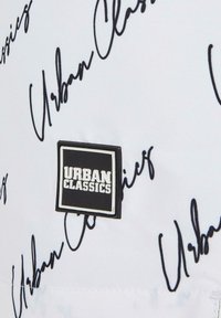 A fehér anyagon merész fekete kézírásos szöveg olvasható: "Urban Classics." Egy téglalap alakú fekete-fehér logó címke van jól látható helyen elhelyezve.
