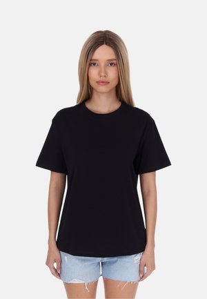 Jonge vrouw met lang, stijl haar, die een effen zwart T-shirt en lichtblauwe gerafelde spijkershorts draagt, staand tegen een effen achtergrond.