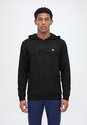 Un homme présente un sweat à capuche noir Adidas avec une poche avant et un pantalon bleu sur un fond blanc uni.