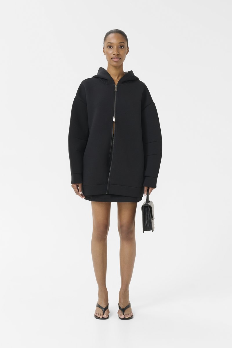 Zwarte oversized hoodie met een rits aan de voorkant, gecombineerd met een zwarte minijurk, een kleine zwarte handtas vasthoudend, en met slippers aan.
