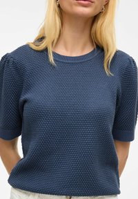 Dziergany sweter z krótkim rękawem w kolorze granatowym z teksturowanym wzorem oraz ściągaczem przy dekolcie i mankietach. Okrągły kształt o luźnym kroju.