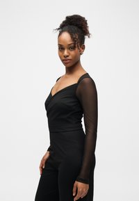 Jeune femme aux cheveux bouclés attachés en chignon haut, portant une tenue noire ajustée avec des manches longues transparentes, debout devant un fond blanc.