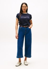 Granatowa koszulka z białym obszyciem z logo "TOMMY HILFIGER", zestawiona z szerokimi, niebieskimi dżinsami i białymi butami ze złotymi akcentami.