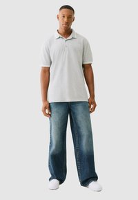 Grijze polo met kraag en logo-detail, gecombineerd met wijde blauwe spijkerbroek en witte sneakers. Soepele stof, relaxte pasvorm.