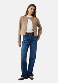 Beige Wildledercropped-Jacke mit rundem Halsausschnitt, getragen über einem weißen, gerippten Top und hoch taillierten blauen Jeans. Ergänzt durch braune Schuhe und einen Gürtel.