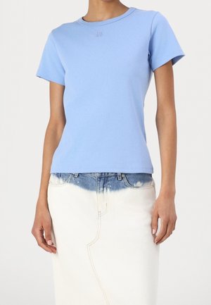 Kvinde iført en tætsiddende lyseblå kortærmet skjorte og hvid nederdel med denimblå topsektion, stående mod en ensfarvet baggrund.