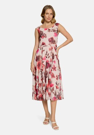 Femme debout, main sur la hanche, portant une robe midi sans manches à imprimé floral rose et marron et des talons blancs ouverts sur un fond uni.