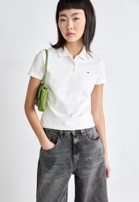 Polo shirt blanc avec un col à trois boutons, petit logo sur la poitrine, associé à un jean noir délavé et ample, et un petit sac vert.