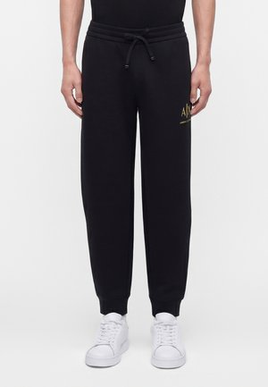 Mann trägt schwarze Jogginghosen mit Kordelzug und goldenem A|X Armani Exchange-Logo nahe der Tasche, kombiniert mit weißen Sneakers vor einfachem Hintergrund.