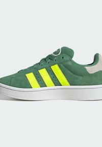 Zapatilla de deporte verde de ante con tres franjas amarillas brillantes, suela blanca texturizada y acentos grises en la parte trasera y en la lengua. Diseño atlético clásico.