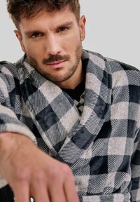 Bata de cuadros gris y azul marino hecha de tela suave, con un cuello estilo chal y textura blanda, con una mano extendiéndose hacia el espectador.