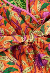 Tissu orange vif avec un motif de feuilles et de baies en vert et violet, accompagné d'un lien rose et de surpiqûres décoratives. Surface texturée.