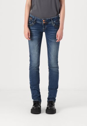 Aangesloten donkerblauwe spijkerbroek van denim met een lichte vervaging. Voorzien van een sluiting met twee knopen, voorzakken en een taps toelopende pijp. Er worden zwarte schoenen gedragen.