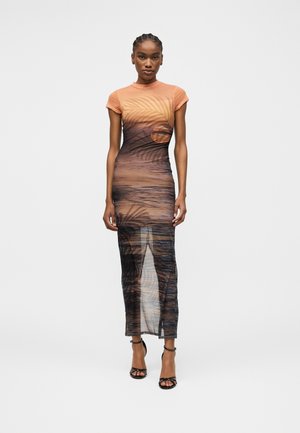 Žena v priliehavej maxi šaty s krátkym rukávom s potlačou západu slnka a paliem, v kombinácii s čiernymi sandálmi na vysokom opätku s remienkami.