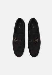 Mocassins en daim noir avec une semelle plate, dotées d'un accent en forme d'ancre dorée et de détails tissés à l'avant. Texture lisse et design classique.