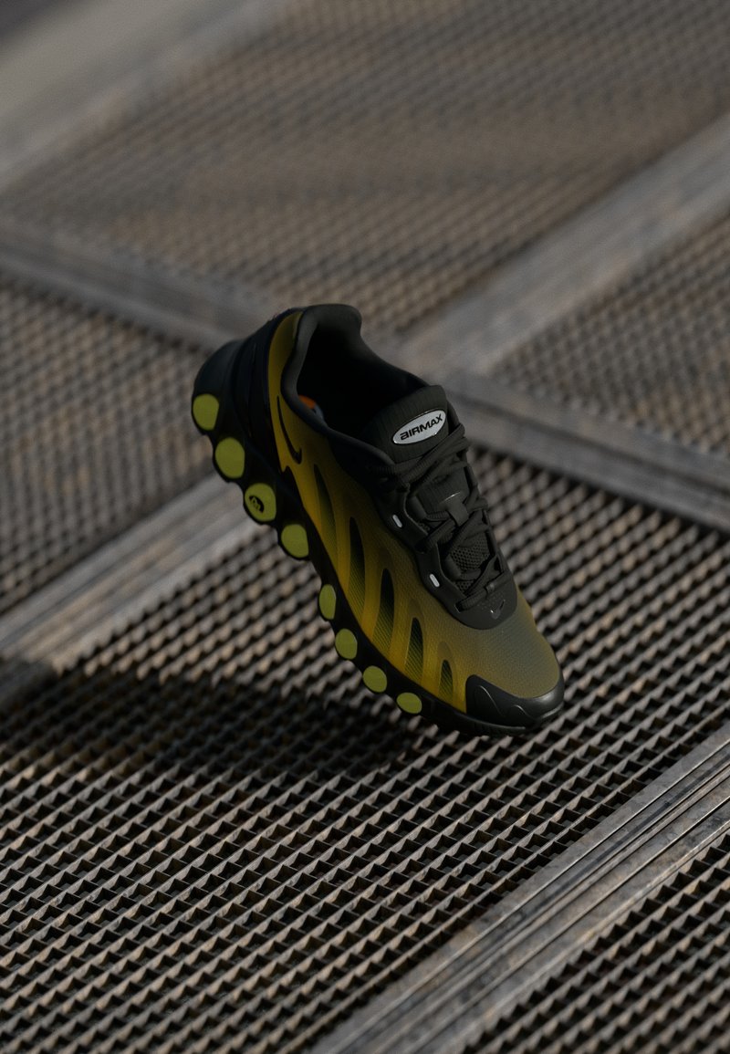 Crna i žuta atletska cipela s teksturiranim gornjim dijelom, mrežastim dizajnom i karakterističnom mjehurićastom srednjom potplatu s zelenim akcentima. Nike brendiranje vidljivo.