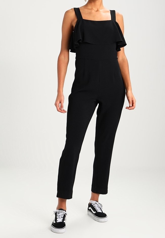 Czarny kombinezon z szerokimi ramiączkami, kwadratowym dekoltem i falbaniastym wykończeniem. Dopasowane nogawki, gładka struktura, noszony z czarno-białymi trampkami.