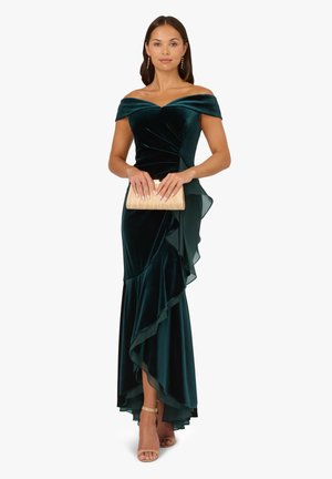 Abito in velluto verde smeraldo con scollo off-the-shoulder, orlo asimmetrico arricciato e dettagli trasparenti. Modella con una pochette dorata.