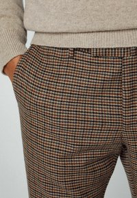 Detailní záběr osoby v béžovém svetru a hnědých kostkovaných kalhotách, s jednou rukou v kapse.
