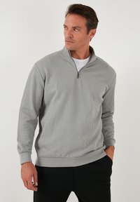 Light gray zip-up pullover made of textured fabric, featuring a stand collar and ribbed cuffs, paired with black pants.