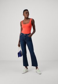 Felrood bodysuit, donkerblauwe jeans met uitlopende pijpen, witte sneakers met groene accenten, en een kleine blauwe handtas.