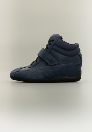 Baskets montantes Puma bleu foncé avec lacets noirs, une bride en Velcro, un col de cheville rembourré et une semelle noire sur fond gris clair.