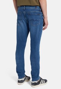 Jeans di denim in un lavaggio blu medio con una vestibilità slim, dotati di due tasche posteriori e dettagli di cucitura sottili. Indossati con sneaker scure.