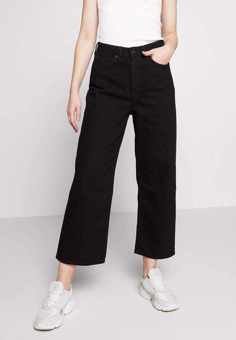 Schwarze weite Jeans aus Denim mit hoher Taille, fünf Taschen-Design und dezenten Nahtdetails. Tragen mit weißen Sneakers.