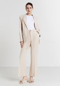 Donna in piedi con indosso pantaloni larghi beige e giacca coordinata sopra una maglietta bianca, accessoriate con anelli dorati e tacchi decorati con fiori.